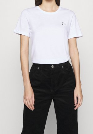 Woman wearing a white T-shirt with a small black smiley face on the left chest and black corduroy pants.