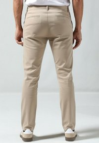 Beige enganliegende Hose mit Gesäßtaschen, glatter Textur, mittelhohem Bund und schlanker Silhouette, kombiniert mit weißen und beigen Sneakern.