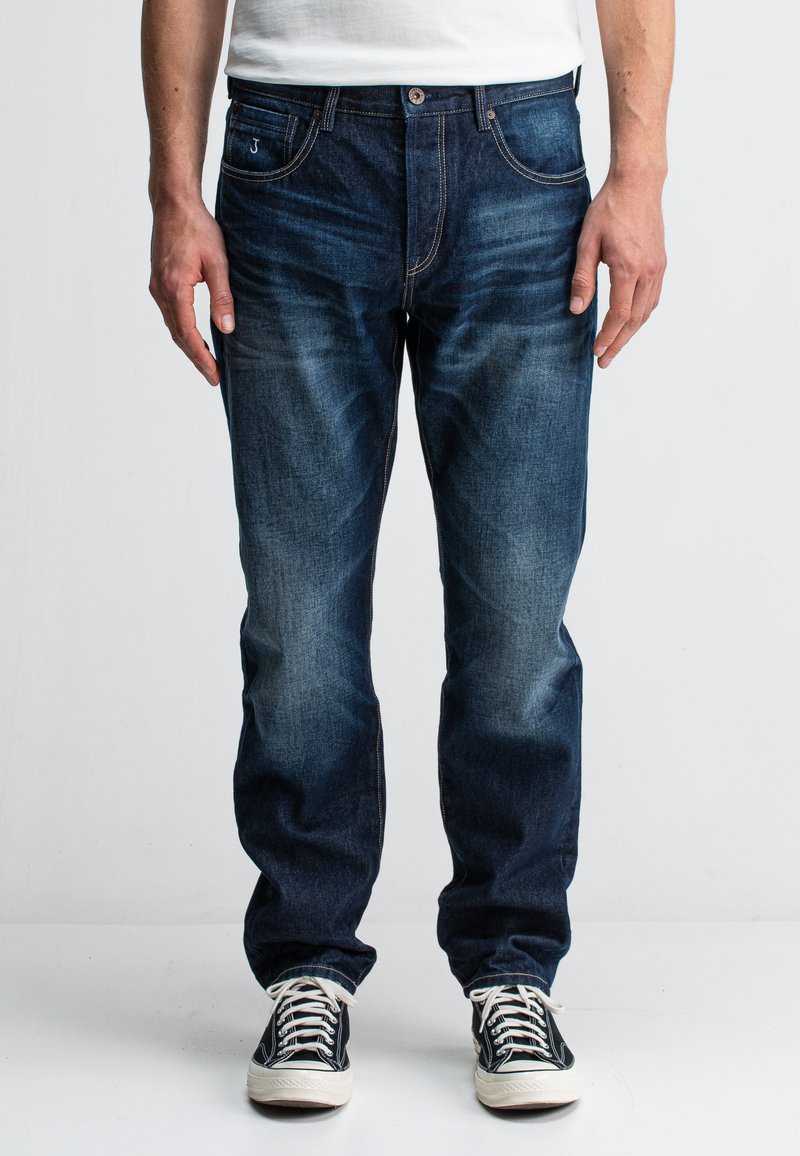 Donkerblauwe denim jeans met een rechte pasvorm, voorzien van vijf zakken, contrasterende stiksels en een vervaagde textuur. Gecombineerd met zwarte sneakers.