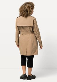 Gabardina color beige con un diseño de doble botonadura, hombreras, una cintura ceñida con cinturón y una clásica abertura trasera, combinada con pantalones negros tipo cropped.