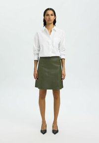 Biała koszula z guzikami, z kieszonką na piersi, połączona z zieloną skórzaną spódnicą w kształcie litery A i czarnymi szpilkami na prostej tle.