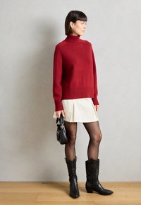 Rød strikket turtleneck genser med ribbestrikket kant, kombinert med en kremfarget plissert skjørt, svarte strømpebukser og svarte cowboy-støvler. En liten svart veske er lagt til.
