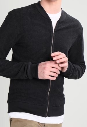 Zwarte bomberjack met ritssluiting, gemaakt van een zachte, textuurstof, met ribgebreide manchetten en zoom, en een zilveren rits. Heeft een ronde halslijn over een wit shirt.