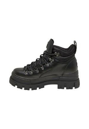Person trägt ein blau gestreiftes Hemd, schwarzen Leder-Midirock und schwarze klobige Schnürstiefel und steht auf einem sonnenbeschienenen Gehweg in der Nähe eines Bordsteins.
