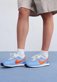 Sneaker sportive azzurro chiaro con accenti arancioni, caratterizzati da una parte superiore testurizzata e lacci bianchi. Suola in gomma con battistrada nero per una buona aderenza.