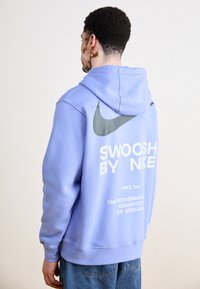 Light blue hoodie with gray Nike swoosh logo on the back. It features white text reading "SWOOSH BY NIKE" and a kangaroo pocket.