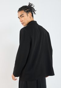 Black tailored jacket with a pointed collar, long sleeves, and a straight fit. Smooth texture and minimal stitching details.