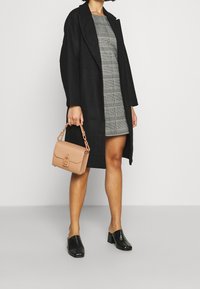 Černý dlouhý kabát přes kárované šaty, s světle hnědou kabelkou se zapínáním a černými texturovanými muly s robustním podpatkem.