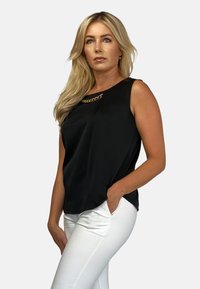 Czarny top bez rękawów o teksturowanej tkaninie z złotym łańcuchem jako akcent przy dekolcie. W połączeniu z białymi dopasowanymi spodniami.