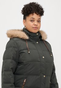 Tumed roheline puff jope, millel on beeži karvaga vooderdatud kapuuts, kuldsete detailide ja lukuga taskud. Liistudega disain ja reguleeritavad nöörid.