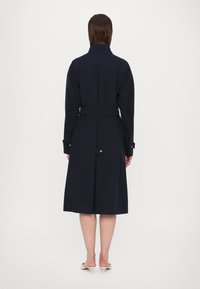 Tumesinine trench coat, millel on vöödiga talje, pikad varrukad ja taga liikuv ventilatsioon. Sile kude, põlvepikkune disain, metallnupud varrukatel.
