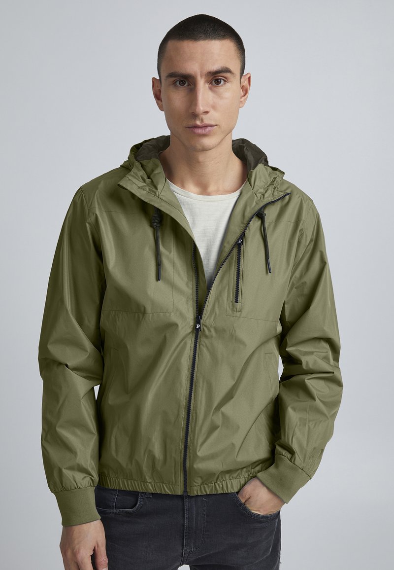 Giacca leggera verde oliva con cappuccio, zip frontale, polsini elastici e due tasche sul petto. Texture liscia e design minimale.