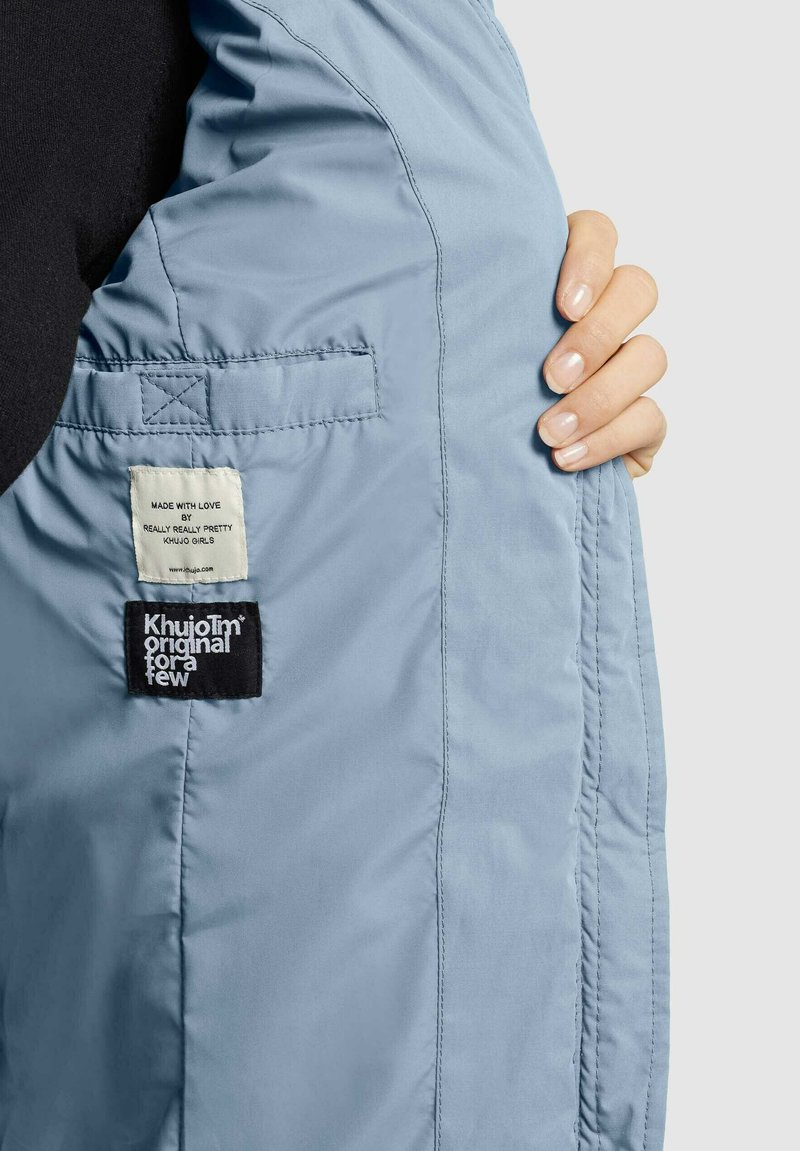 Helleblaue Jacke mit glatter Textur, Innenetiketten und auffälligen Steppdetails. Der Stoff ist leicht und atmungsaktiv.