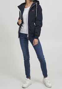 Granatowa kurtka przeciwwiatrowa z kapturem, białym zamkiem błyskawicznym i naszywką z logo. Połączona z dopasowanymi niebieskimi dżinsami i białymi trampkami. Gładka faktura.