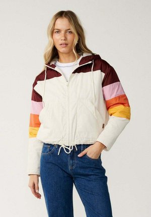 Femme aux longs cheveux blonds portant une veste crème avec des rayures bordeaux, roses, orange et jaunes, et un jean bleu, debout avec une main dans la poche.