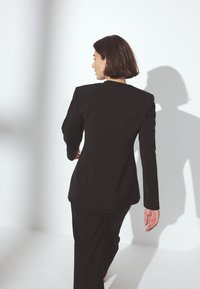 Svart skräddarsydd blazer med strukturerade axlar och långa ärmar, tillsammans med matchande byxor, mot en ljus bakgrund.