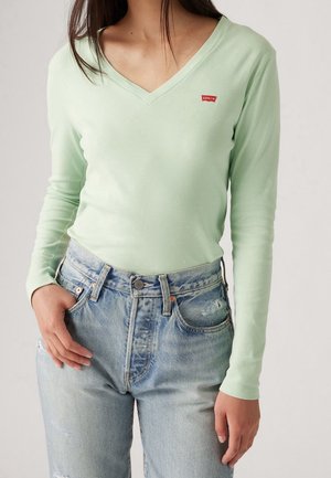 Vrouw draagt een lichtgroen T-shirt met lange mouwen en V-hals en een hooggetailleerde lichtblauwe spijkerbroek met lichte slijtage.