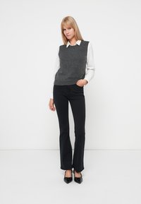 Pulover gri închis cu decolteu în V, purtat peste o cămașă albă cu guler. Pantaloni negri evazați și pantofi negri completează ținuta.