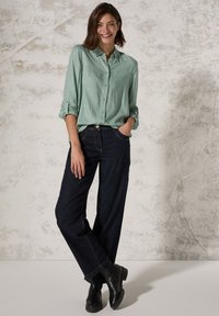 Camicia a righe verde chiaro a maniche lunghe con taschino sul petto, abbinata a jeans larghi blu scuro e stivaletti neri. Sfondo neutro con texture.