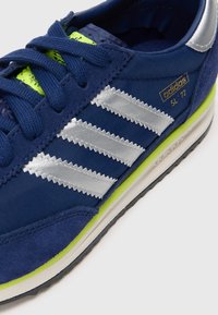 Modré tenisky Adidas SL '72 so striebornými pruhmi, neonovo zelenými akcentmi a tmavomodrými šnúrkami na bielom pozadí.
