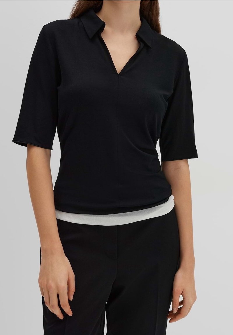 Schwarzes Kurzarmshirt mit Kragen und dezentem Glanz. Es hat eine taillierte Passform und eine glatte Textur, getragen zu schwarzen Hosen.