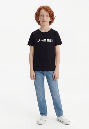 Rude dziecko stoi przodem, ubrane w czarną koszulkę z logo Messiego, jasnoniebieskie dżinsy i białe trampki na białym tle.