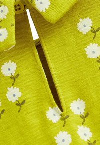 Primo piano di un tessuto giallo-verde con stampa di fiori bianchi e un piccolo bottone in alto, che mostra una fessura sotto il bottone.