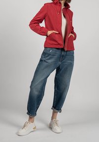 Rød letvægtsjakke med lynlås, elastiske manchetter, båret over en cremefarvet top, parret med løse blå jeans og hvide sneakers.