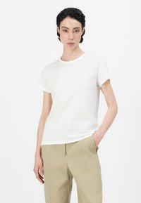 Weißes T-Shirt aus Baumwolle mit kurzen Ärmeln, rundem Halsausschnitt und kleinem Blumenemblem auf der linken Brust. Kombiniert mit beigen Hosen.