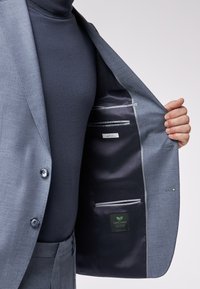 Giacca di suit azzurra chiara con una texture liscia, caratterizzata da un rivestimento scuro, tasche interne e un'etichetta con la scritta "SALUTANDO L'EVOLUZIONE SOSTENIBILE."