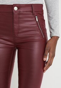 Primer plano de una persona que lleva pantalones de cuero burdeos con un bolsillo lateral con cremallera, un botón plateado y una camiseta de manga larga gris.