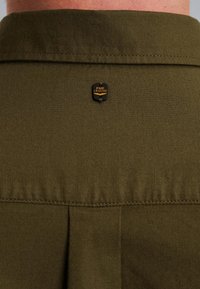 Camicia verde oliva con colletto punta, caratterizzata da un piccolo distintivo nero e oro PME Legend sul retro e un dettaglio a pieghe all'orlo.
