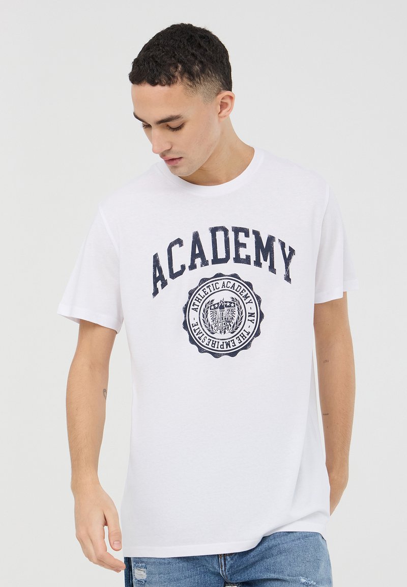 T-shirt bianco in cotone con maniche corte, caratterizzata da una grafica "ACADEMY" blu navy e un design con distintivo circolare sulla parte frontale.
