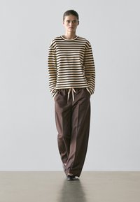 Donna che cammina indossando un top a maniche lunghe beige e nero a righe, pantaloni larghi marroni con coulisse e scarpe nere su uno sfondo uniforme.