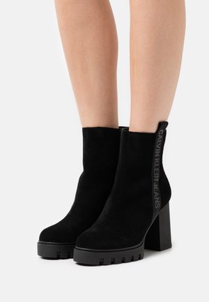 Stivali alla caviglia neri in suede con tacco a blocco massiccio e suola in gomma testurizzata. Presentano un anello laterale con il marchio "CALVIN KLEIN JEANS".