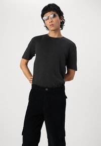 Sort ribbet t-shirt parret med sorte corduroy cargo bukser. Accessories inkluderer rektangulære sølvfarvede solbriller. Modellen står med hænderne på hoften.