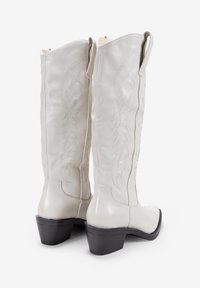 Stivali bianchi in pelle al ginocchio con motivi floreali ricamati, punta quadrata e un tacco bloccato basso nero. Chiusura con zip sul retro.