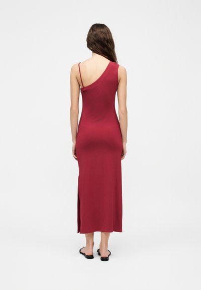 Γυναίκα με μακριά μαλλιά φοράει ένα κόκκινο φόρεμα χωρίς μανίκια, με έναν ώμο και πλαϊνό σκίσιμο, συνδυασμένο με μαύρα, επίπεδα σανδάλια, στέκεται σε μονόχρωμο φόντο.