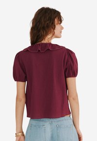 Blusa corta bordeaux a maniche corte con colletto arricciato e tessuto texture. Il retro presenta una vestibilità rilassata e pieghe sottili sulle spalle.