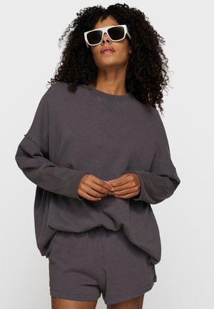 Donna con capelli ricci che indossa occhiali da sole bianchi, felpa oversize scura e pantaloncini coordinati, in piedi davanti a uno sfondo semplice.