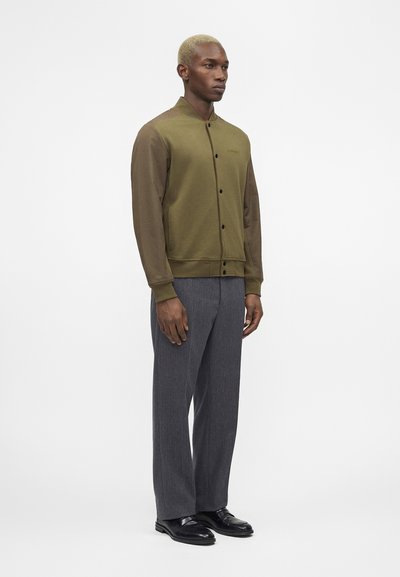 Άνδρας στέκεται απέναντι φορώντας ένα χακί πράσινο κουμπωτό μπουφάν με καφέ μανίκια, γκρι παντελόνι και μαύρα επίσημα παπούτσια σε απλό φόντο.