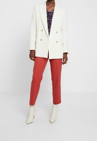 Dubbelknäppt vit kavaj med guldknappar, kombinerad med en mörk blommig topp och röda skräddarsydda byxor, avslutad med krämfärgade spetsiga ankelstövlar.