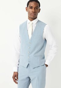 Heledsinise pinstripe'iga vest kuue nööbiga ja taskuga, koos valge pluusi ja kreemjat värvi lipsuga, omades täpset istuvust.