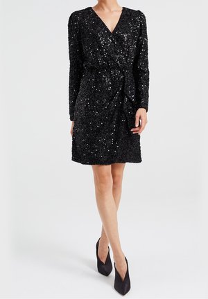 Woman wearing a black long-sleeve sequin dress with a wrap front and black pointed high heels, standing on a white background.