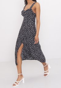 Černé květinové šaty s bílými sedmikr�áskami, volánkovým výstřihem, tenkými ramínky, vysokým rozparkem a lehkým materiálem; nošeny s bílými sandály na podpatku.