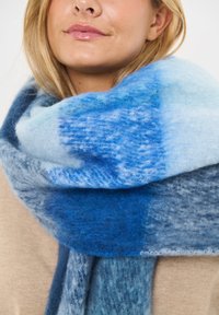 Uldhalstørklæde i forskellige nuancer af blå med et blødt, tekstureret design og farveovergange i gradient. Løst viklet omkring halsen.