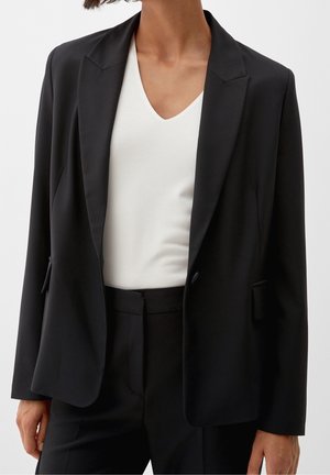 Blazer noir avec un col cranté, deux poches avant et une coupe cintrée, porté sur un haut blanc à col en V. Texture de tissu lisse.