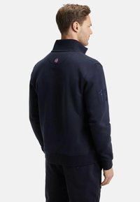 Sweatshirt bleu marine avec col montant, coutures latérales et logo brodé au dos, avec une texture similaire à celle de la polaire.