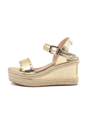 Sandalia tipo cuña metálica dorada con adornos de tachuelas, punta abierta, correa ajustable en el tobillo y plataforma trenzada de yute.