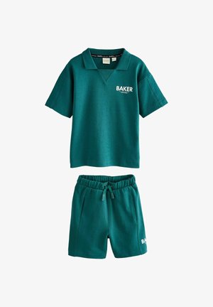 Jongen staat binnen in teal shorts en een polo shirt met witte sneakers en sokken, kijkend naar de zijkant.